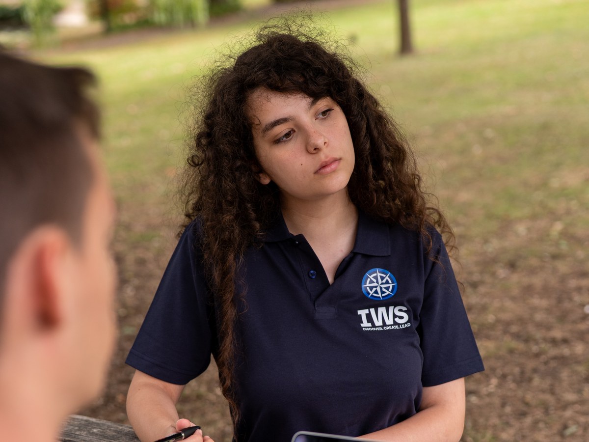 An IWS A Level student in a navy school polo, looking up from her notebook with a pen in her hand, at a table in a park