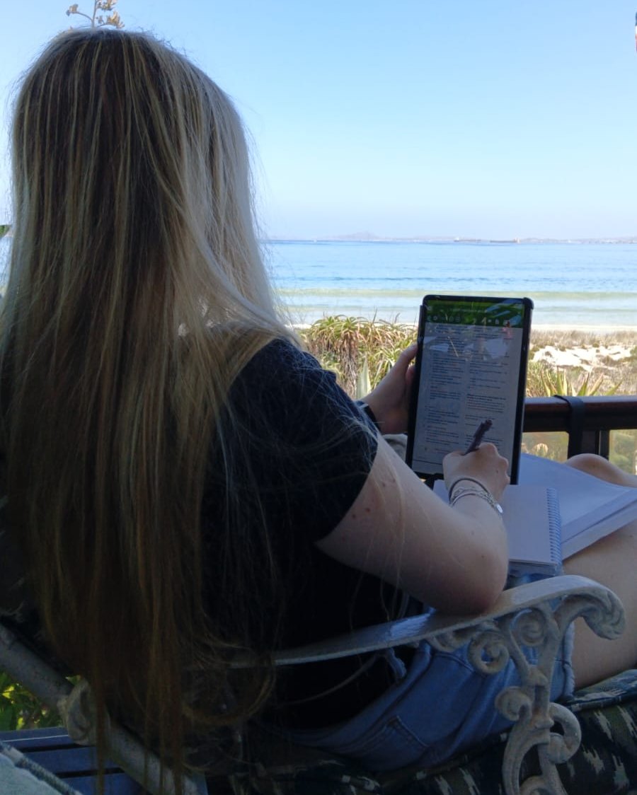 A student with long fair hair, seen from behind, writing on a tablet beside a notebook on a veranda by the sea
