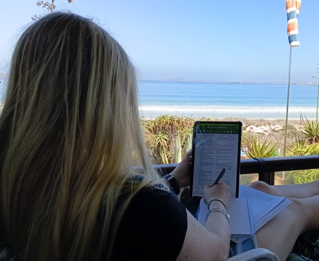 A student with long fair hair, seen from behind, writing on a tablet beside a notebook on a veranda by the sea