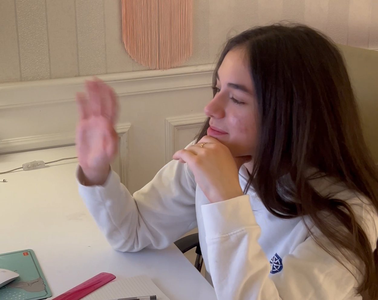 A student in an IWS hoodie at a white desk at home, smiling and lifting a hand