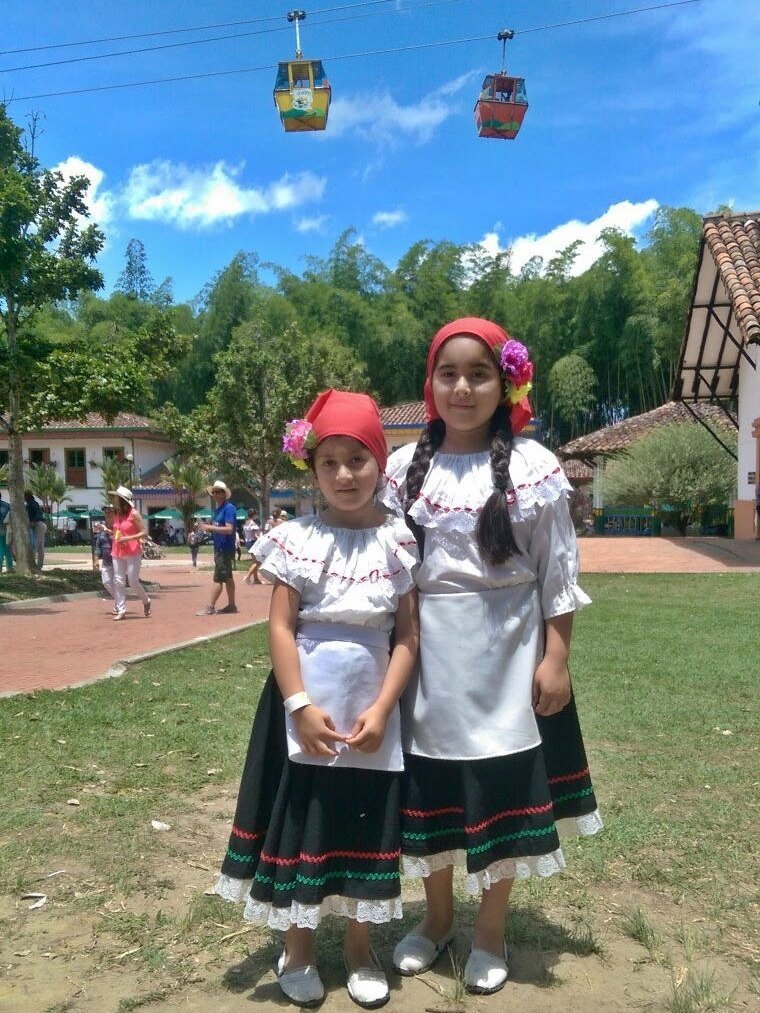Duas meninas com trajes tradicionais colombianos, blusas brancas bordadas, saias pretas com detalhes vermelhos e verdes e lenços vermelhos com flores, em um gramado entre árvores, sob dois teleféricos
