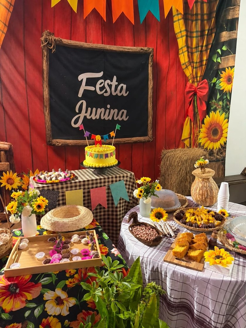 Uma mesa de Festa Junina, com bandeirinhas, uma lousa com as palavras Festa Junina, um bolo amarelo, girassóis e cestas de doces sobre uma toalha xadrez