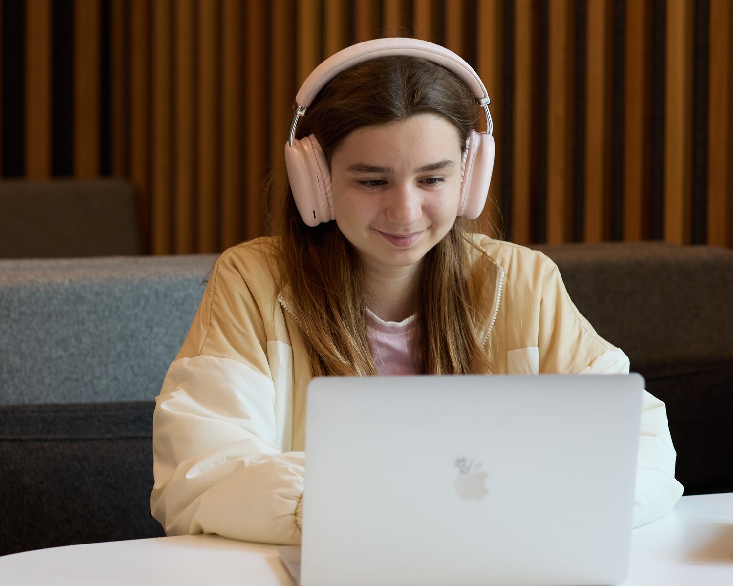 A student in over-ear headphones, smiling at her laptop at a white table