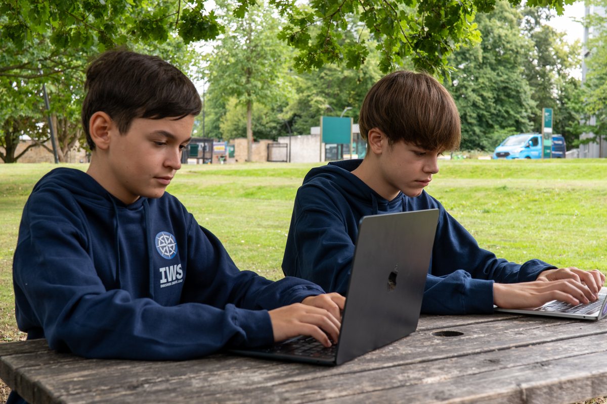 Two IWS students in navy hoodies typing at laptops side by side at a wooden table in a park