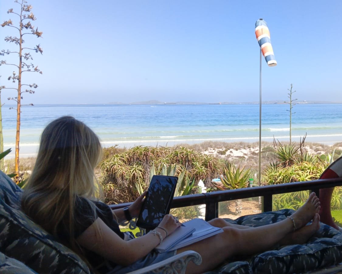 An IWS student, seen from behind, studying from a tablet and a notebook on a veranda by the sea