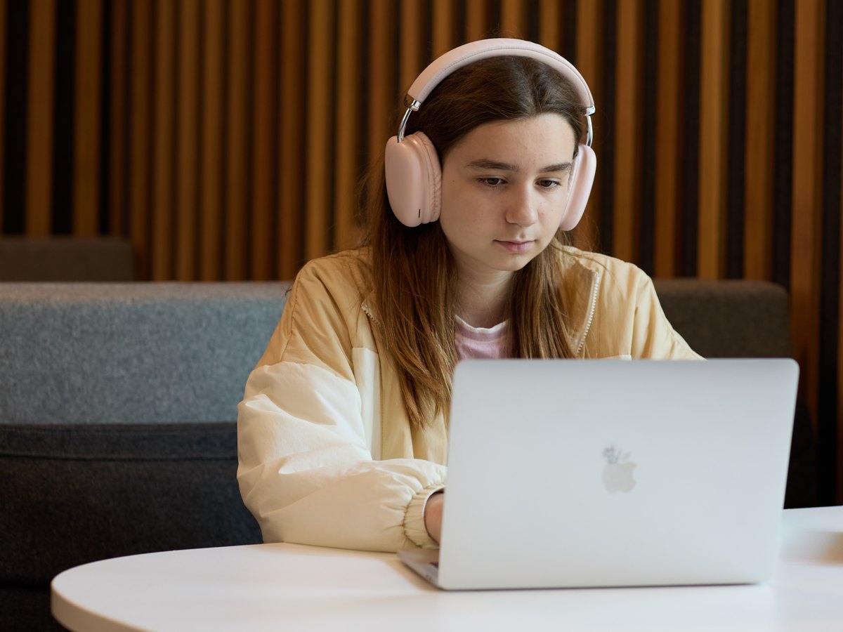 A student in pink headphones and a cream jacket, working at a laptop at a round table