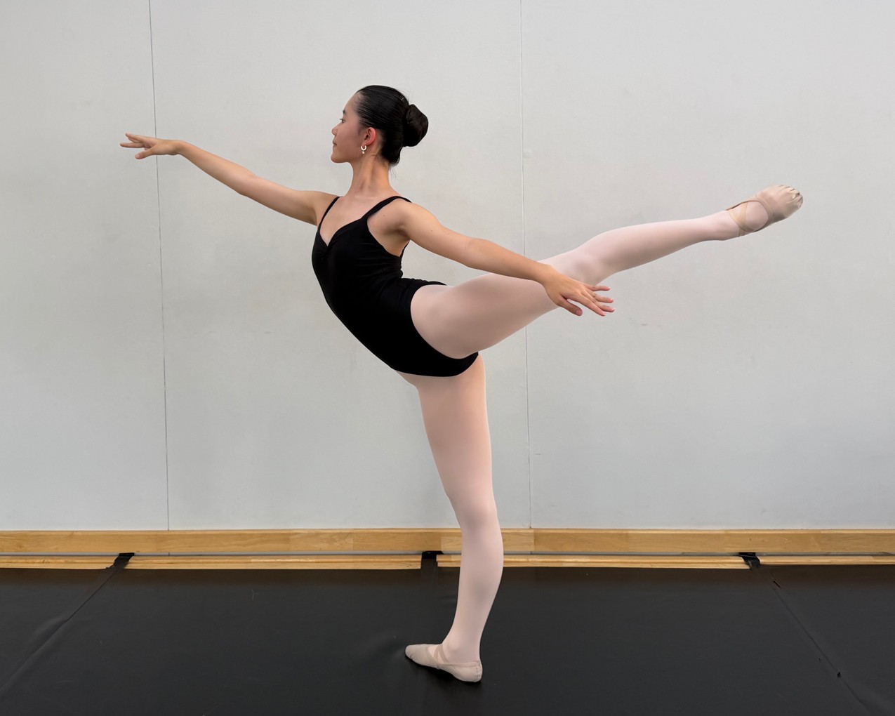 Kei Y., an IWS IGCSE student, in a long arabesque in a bright ballet studio, in a black leotard and pink tights