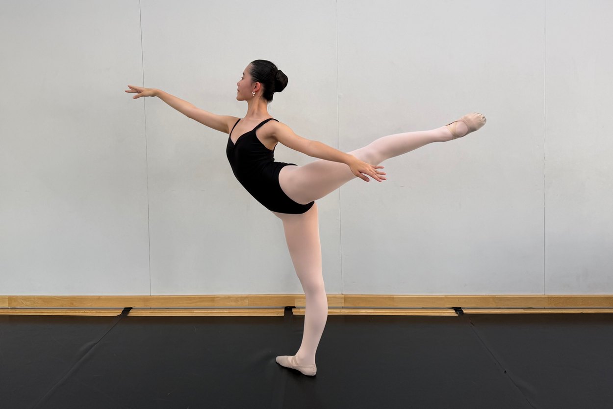 Kei Y., an IWS student, in a long ballet arabesque in a studio, her arms out and one leg raised behind her