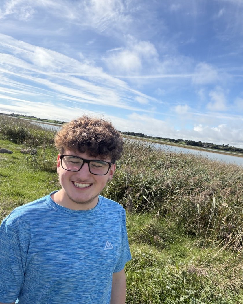 Alfie D., a student with curly hair and glasses, smiling at the camera in front of tall reeds and an estuary under a bright sky