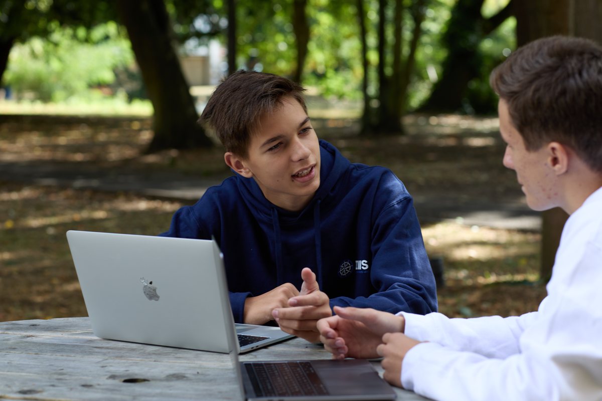 Two IWS students at a round wooden table in a park, each with an open laptop, talking to one another