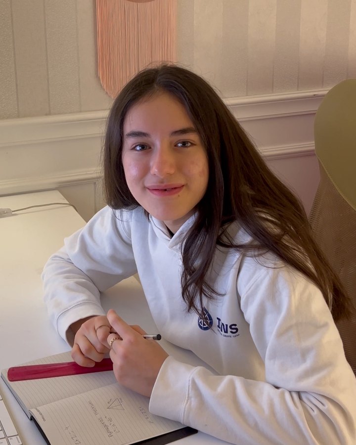 An IWS student at her desk at home, in her IWS hoodie, smiling at the camera with her notebook open
