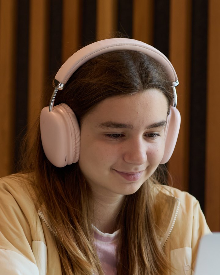 A student in pink headphones and a cream jacket, smiling as she works at her laptop