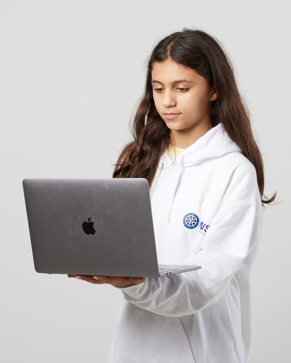 An IWS Primary student in a white school hoodie, looking down at the open laptop she holds, against a plain grey wall