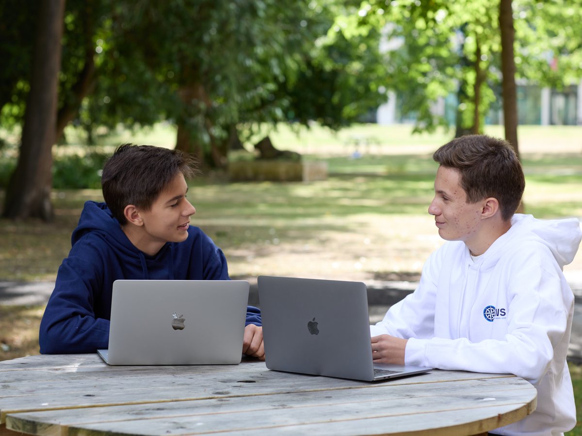 Two IWS students at a round wooden table in a park, each with an open laptop, one turned towards the other