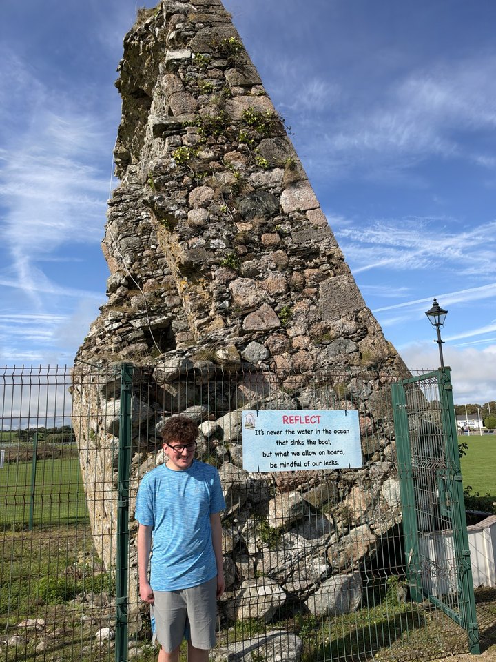 Alfie D., in a blue T-shirt and grey shorts, standing in front of a huge stone ruin that leans sharply, under a blue sky