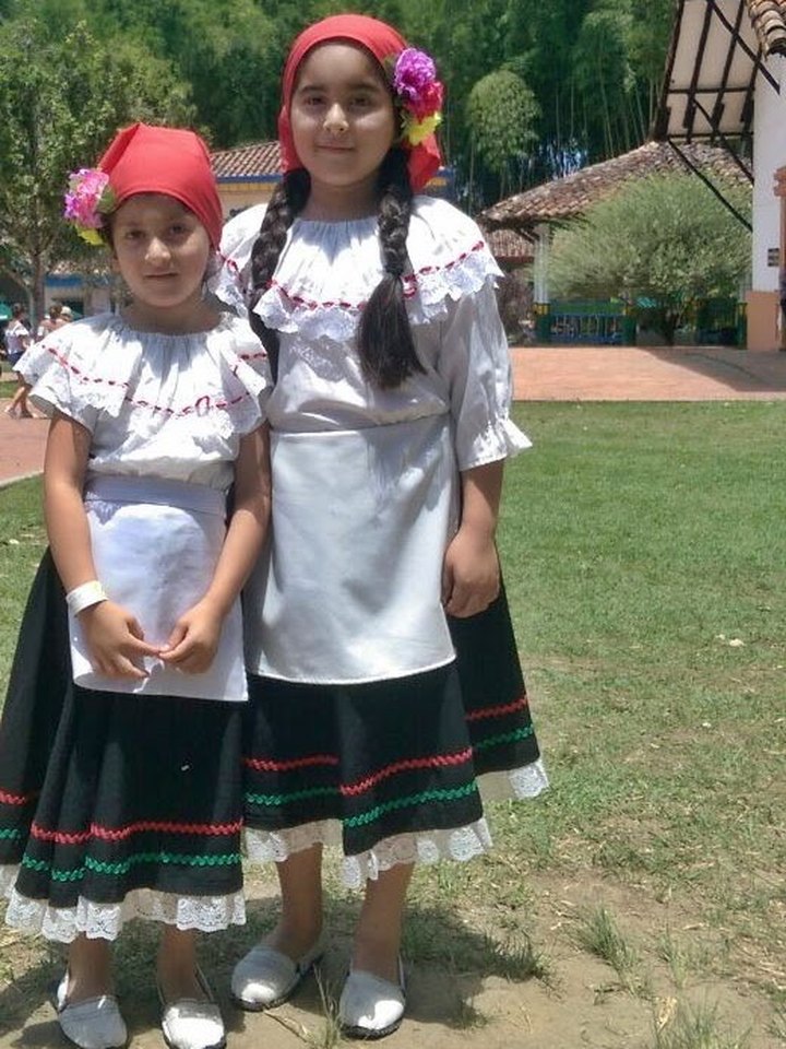 Two girls in traditional Colombian dress, white embroidered blouses, black skirts trimmed in red and green and red headscarves with flowers, standing on a lawn among trees