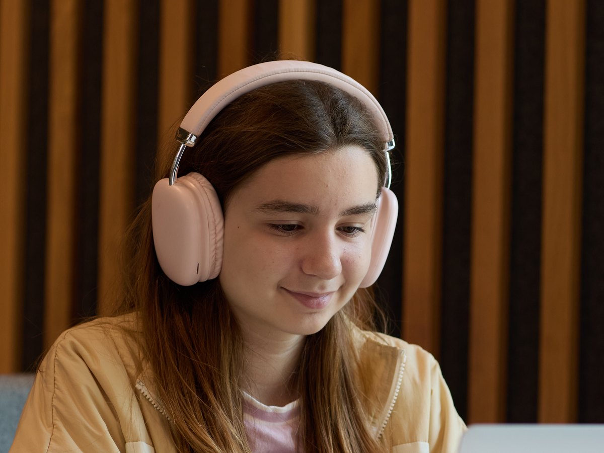 A student in pink headphones and a cream jacket, smiling as she works at her laptop