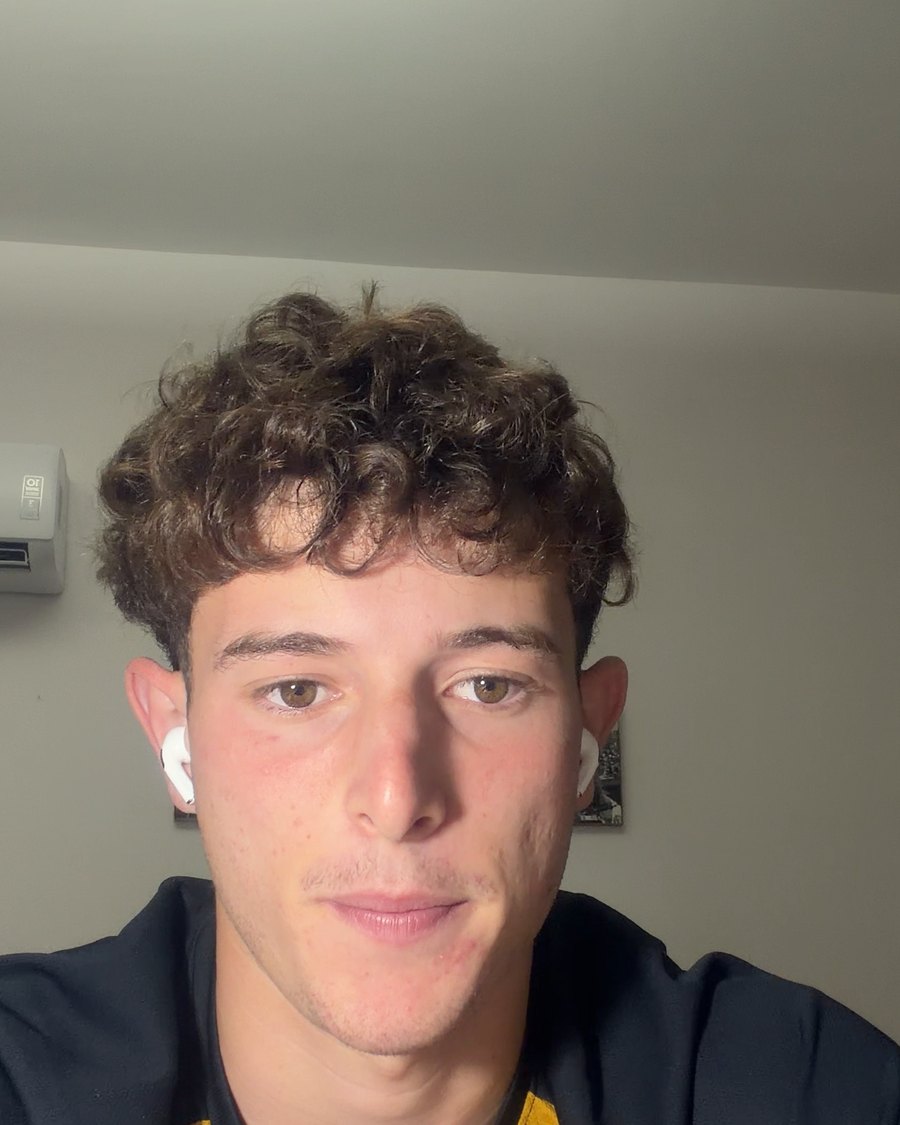 Adrian E., with curly dark hair and a white earbud, looking at the camera