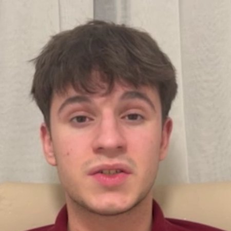 Alexandru R. speaking to the camera in a maroon polo shirt, with pale curtains behind him