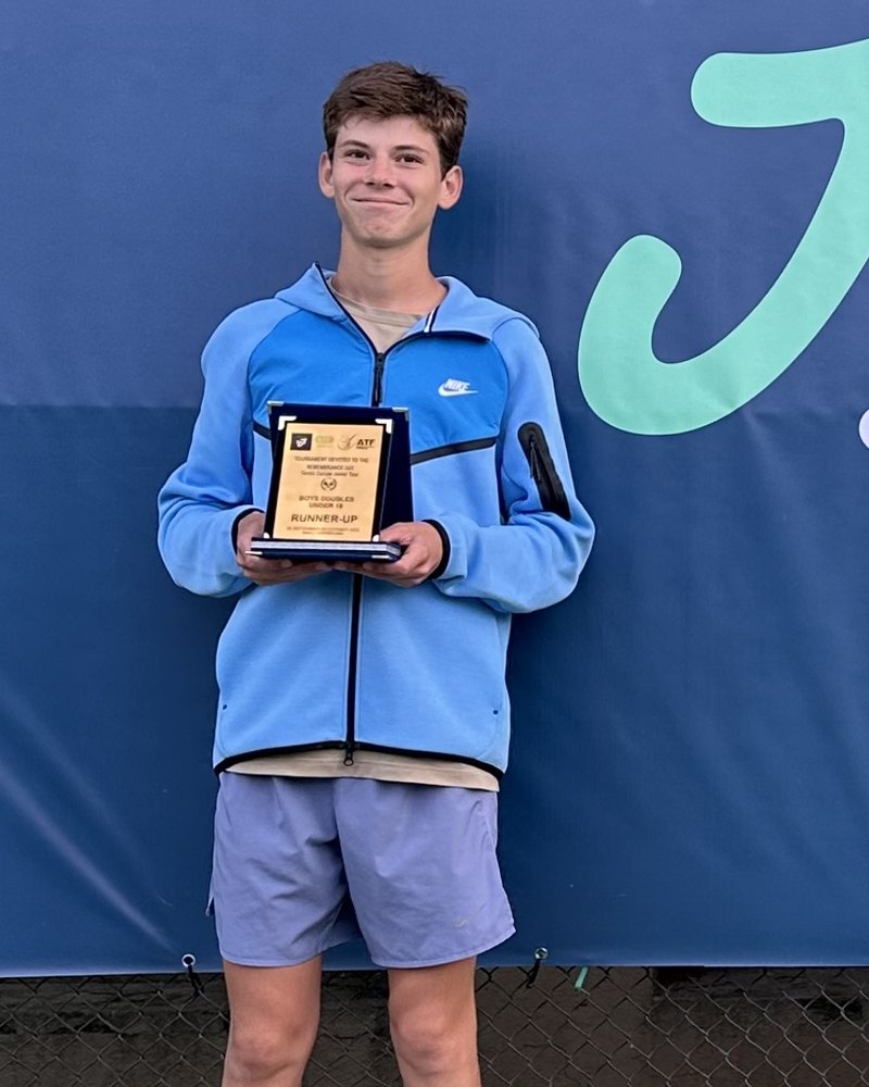 Georgiy G., smiling, in a light-blue jacket and holding a plaque, in front of a blue tournament banner