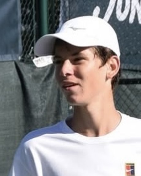 Georgiy G. in a white cap and T-shirt on a tennis court, a racket in his hand, looking away to his left