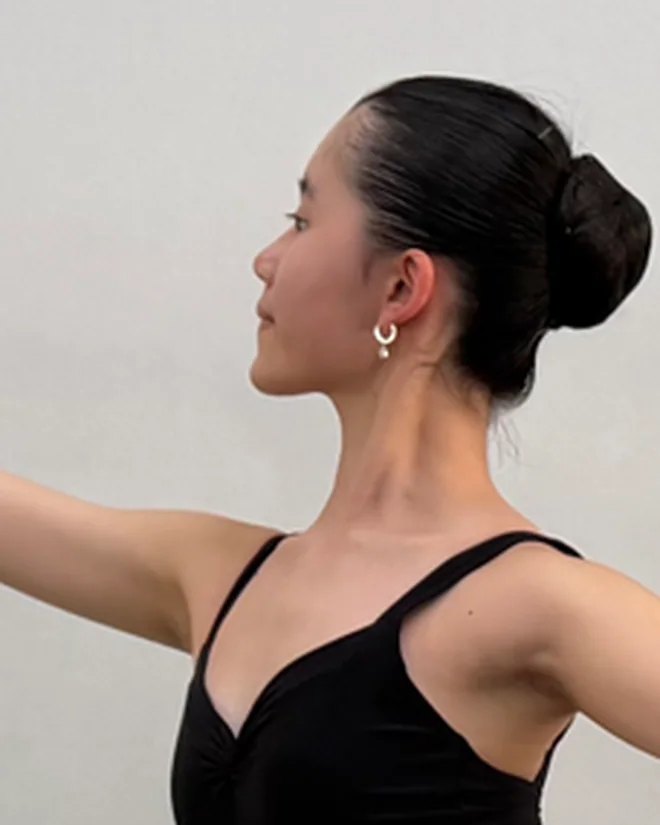 Kei Y. in profile, in a black leotard, with her dark hair in a bun and one arm held out