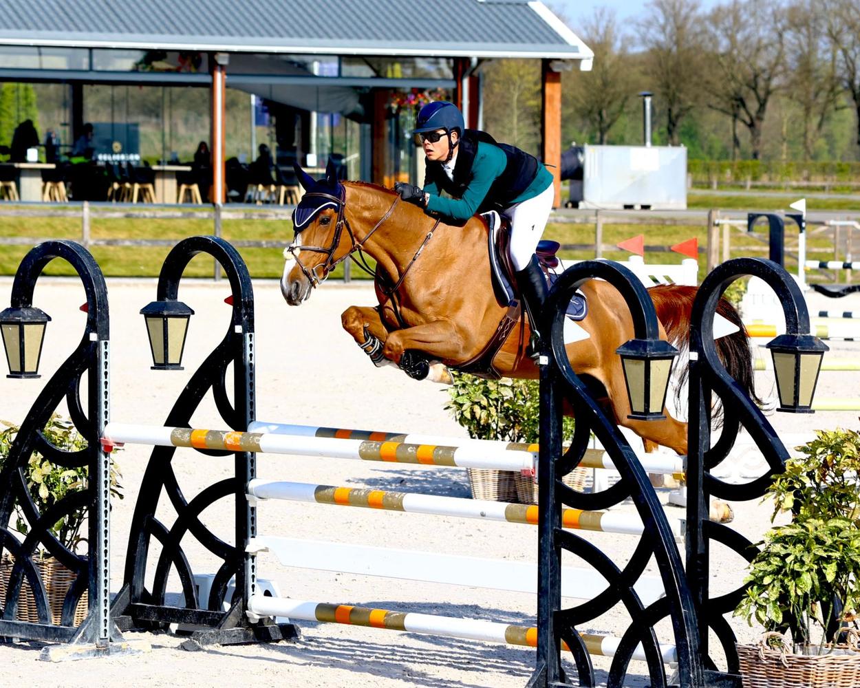 Peter G., in a green jacket and navy helmet, and his chestnut horse clearing a black fence in a show-jumping arena, with a clubhouse behind them