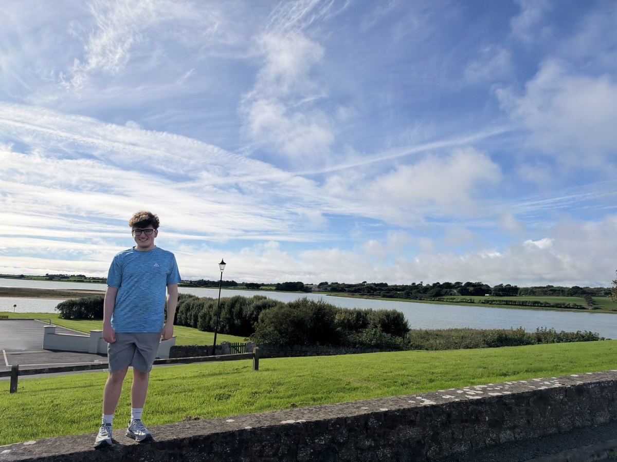 Alfie D., a student in a blue T-shirt, standing small on the left of a wide view of an estuary and green fields under a sky of thin cloud