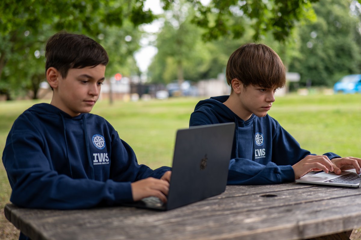 Two IWS students in navy hoodies working at laptops side by side at a wooden table in a park