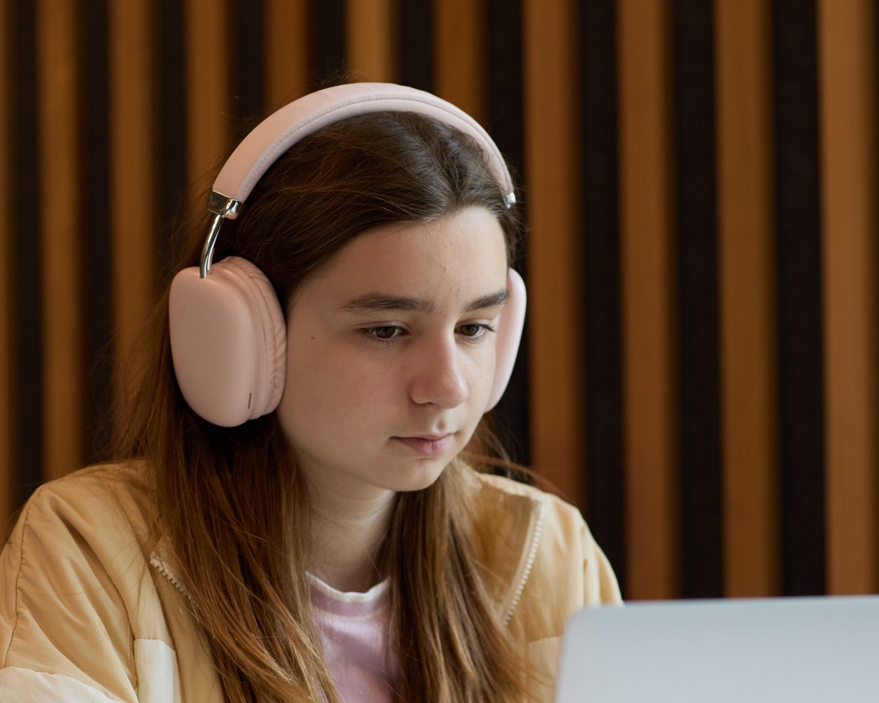 A student in pink headphones looking at a laptop screen, in front of a wall of wooden slats