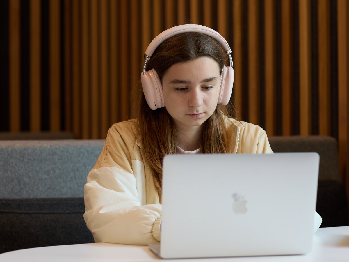 A student in pink headphones and a cream jacket, working at a laptop at a round table